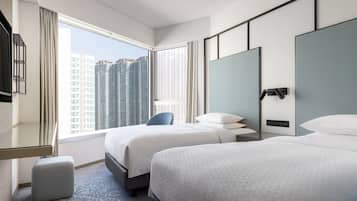 A hotel room with two beds, a large window with a city view, a desk with a chair, and a small ottoman.