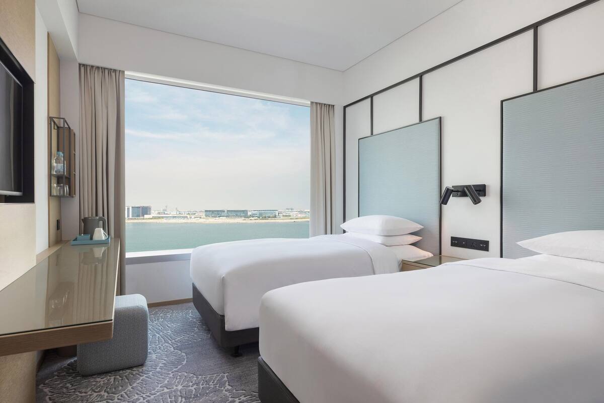 A hotel room with two beds, a desk, a TV, and a large window offering a view of the city.