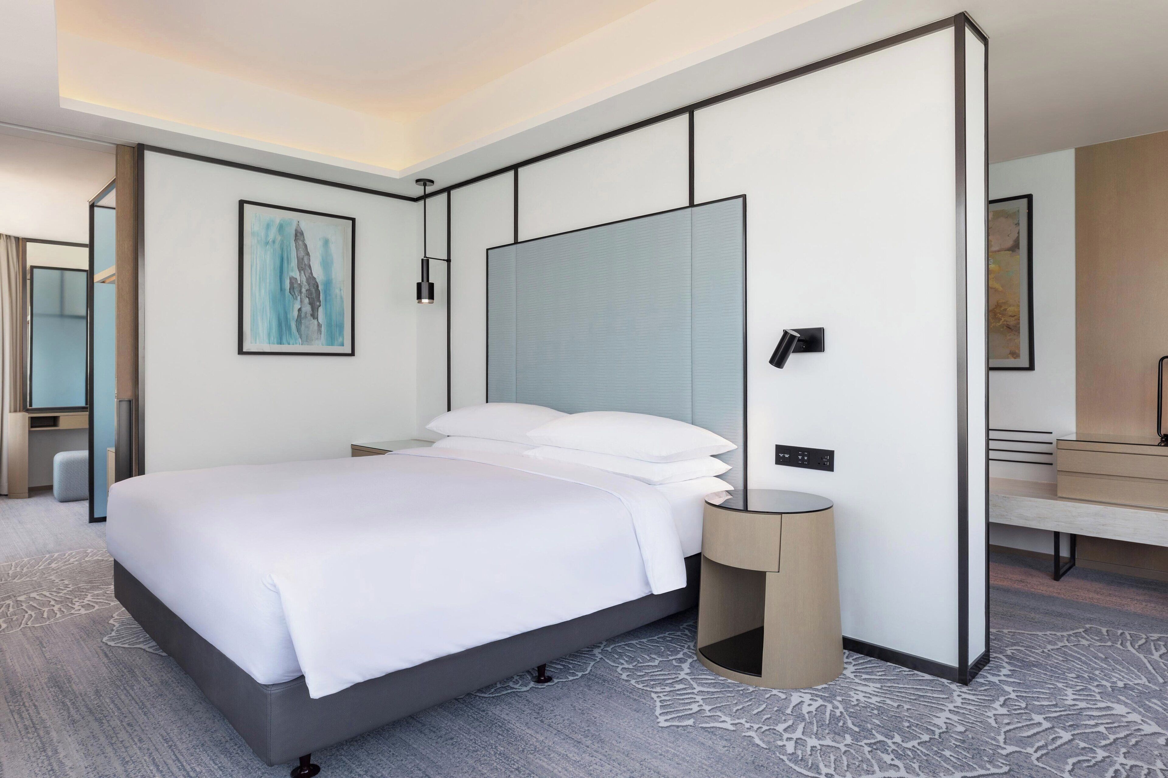 A modern hotel room with a large bed, a bedside table, a nightstand, a mirror, and a wall-mounted artwork.