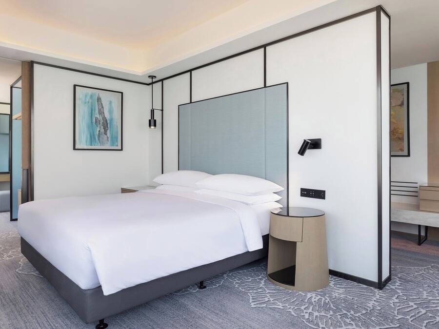 A modern hotel room with a large bed, a bedside table, a nightstand, a mirror, and a wall-mounted artwork.