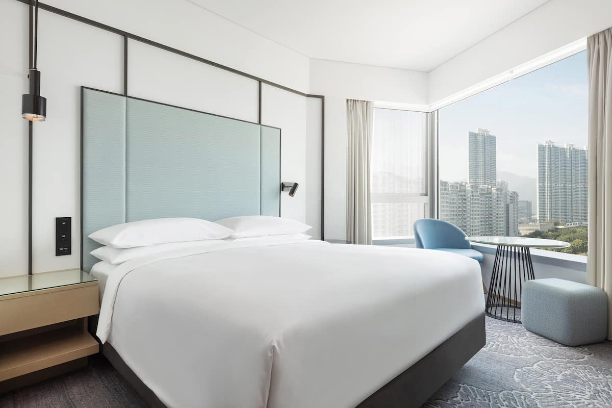 A modern hotel room with a large bed, a glass partition headboard, a small table, and a view of high-rise buildings.