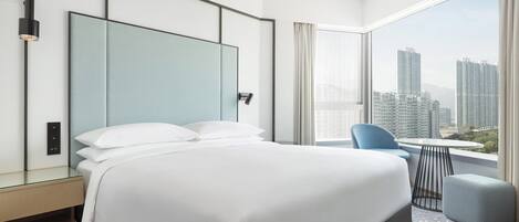 A modern hotel room with a large bed, a glass partition headboard, a small table, and a view of high-rise buildings.