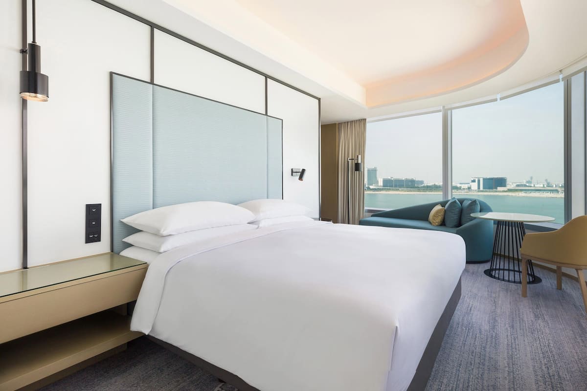 A modern hotel room with a large bed, a sofa, a small table, and a view of the city through large windows.