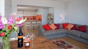 Ein modernes Wohnzimmer mit grauem Sofa, roten und bunten Kissen, einem Holztisch, einer Vase mit Blumen, einer Weinflasche und Einmachgläsern.