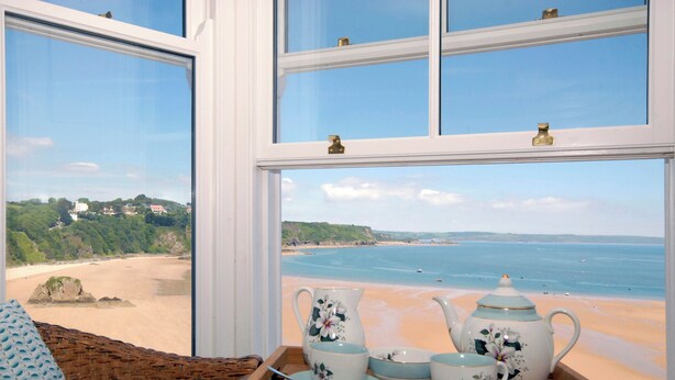 Blick aus dem Fenster auf einen Strand, mit einer Teekanne und einer Kaffeetasse auf einem Holztisch.