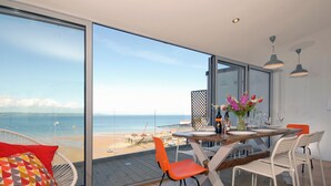 Ein Essbereich mit Blick auf den Strand, ein Tisch gedeckt mit Tellern und Gläsern sowie eine Vase mit Blumen.