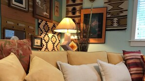 A living room with a beige sofa, multiple cushions, a knitted throw, and wall decorations including framed pictures and a woven tapestry. - Quiet, Stand-alone Country Granny Cottage near the Village, (Arroyo Grande)