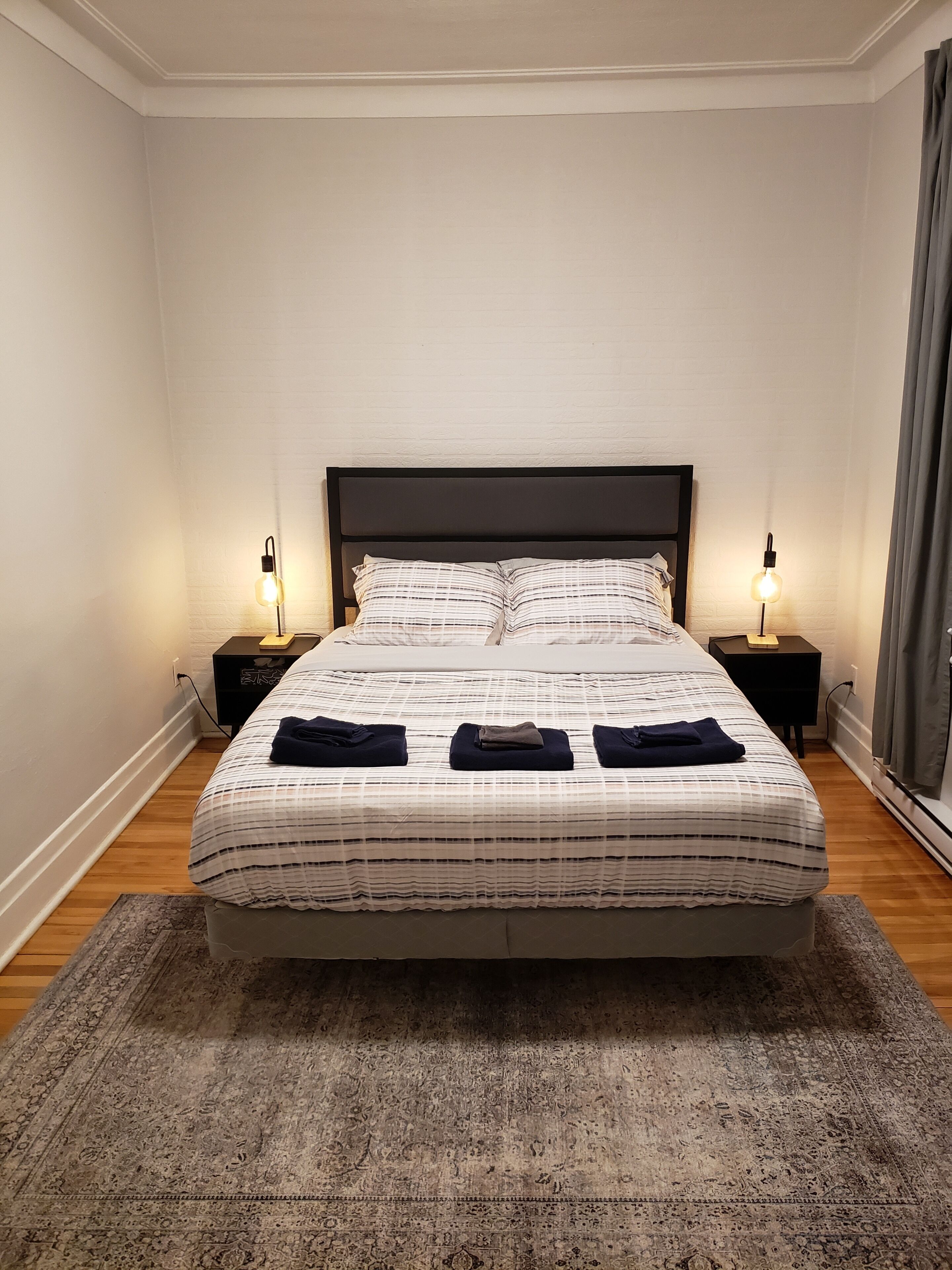 Un lit bien fait, recouvert d’une couvre-lit à rayures, deux tables de chevet avec des lampes et un tapis à motifs sur un plancher en bois.