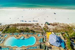 An aerial view of a beach resort with a large pool, sun loungers, and a sandy beach.