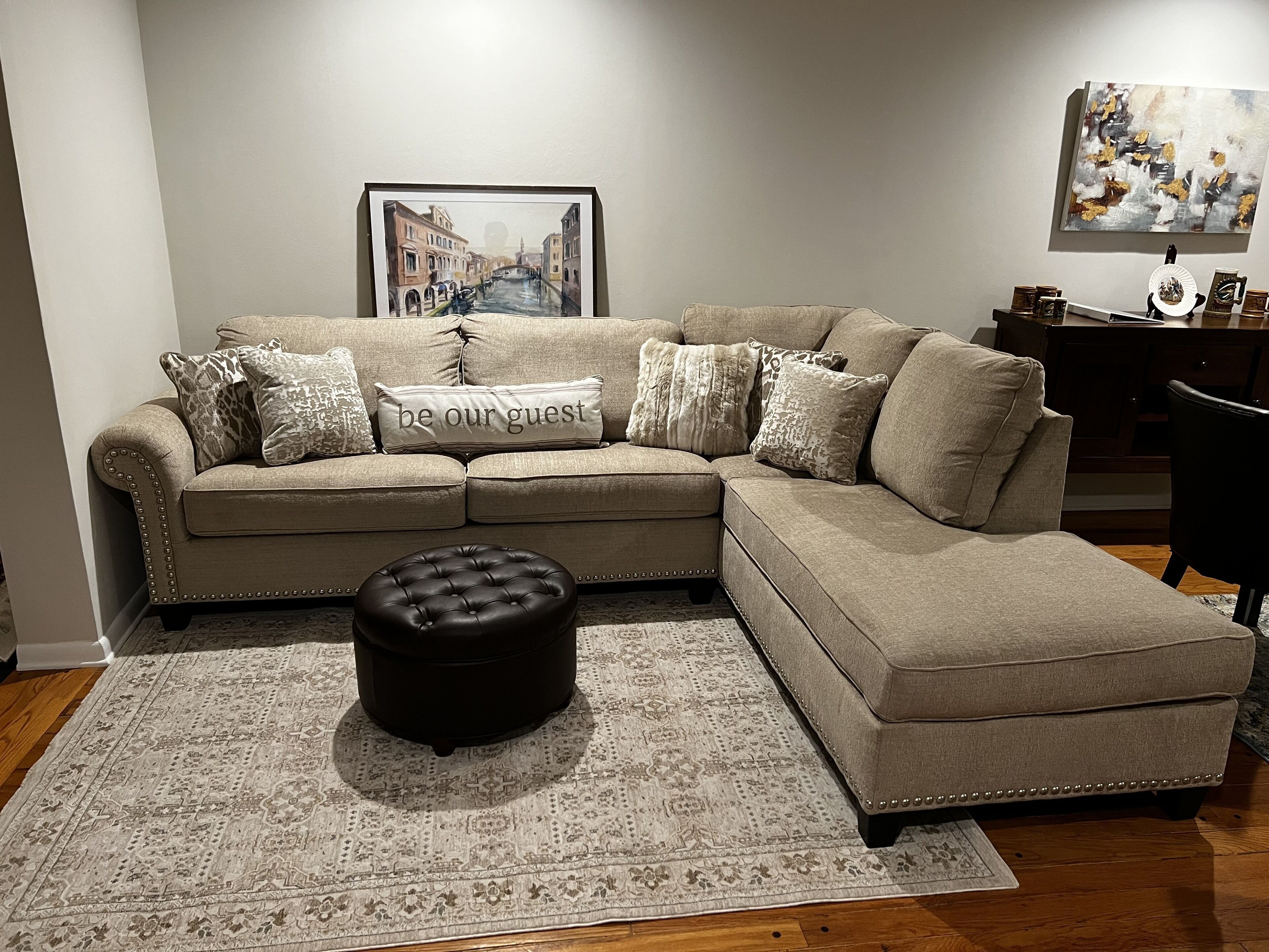 Un canapé d’angle beige agrémenté d’un coussin orné de la mention « soyez les bienvenus », un pouf noir et un tapis à motifs posé sur un plancher en bois.