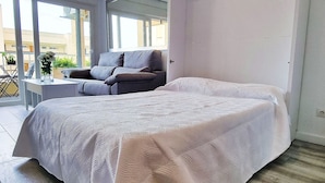 Un dormitorio con una cama grande, un sofá gris, una mesita de noche con un florero y vistas al exterior a través de amplias ventanas.