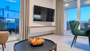 Ein modernes Wohnzimmer mit einem an der Wand befestigten Flachbildfernseher, einem runden Couchtisch mit Obstschale, einem grünen Sessel und Blick auf ein Gebäude durch große Fenster.