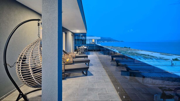 Una terraza moderna al aire libre con vista al mar, que incluye una silla colgante, un área de descanso y una barandilla de vidrio.