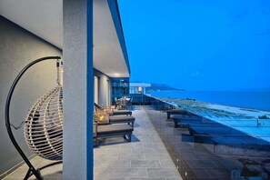 Una terraza moderna al aire libre con vista al mar, que incluye una silla colgante, un área de descanso y una barandilla de vidrio.