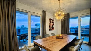 Un comedor con una mesa de madera, sillas tapizadas en gris, una araña y vistas de la ciudad a través de amplias ventanas.