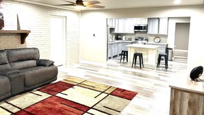 A modern kitchen with white cabinets, a central island, and a dining area with a gray sofa and patterned rug.