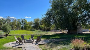 Une terrasse dotée de fauteuils Adirondack et d’un foyer, entourée d’un chemin de pierre et d’une zone herbeuse agrémentée d’une clôture et d’arbres.