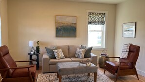 Un salon comprenant un canapé, deux fauteuils, une table basse, une lampe, un tableau et une fenêtre ornée d’un rideau à motifs.