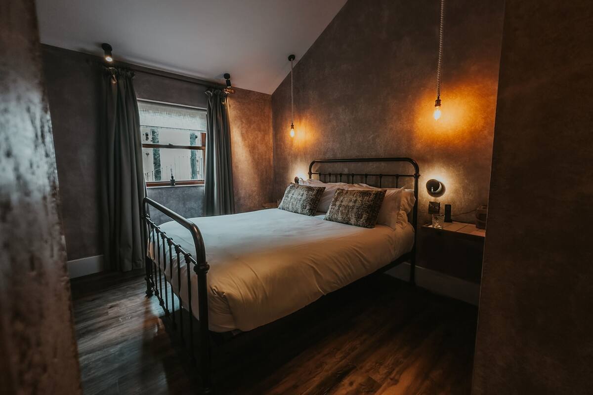 A bedroom with a metal bed frame, a bedside table with a lamp, and a window with curtains.
