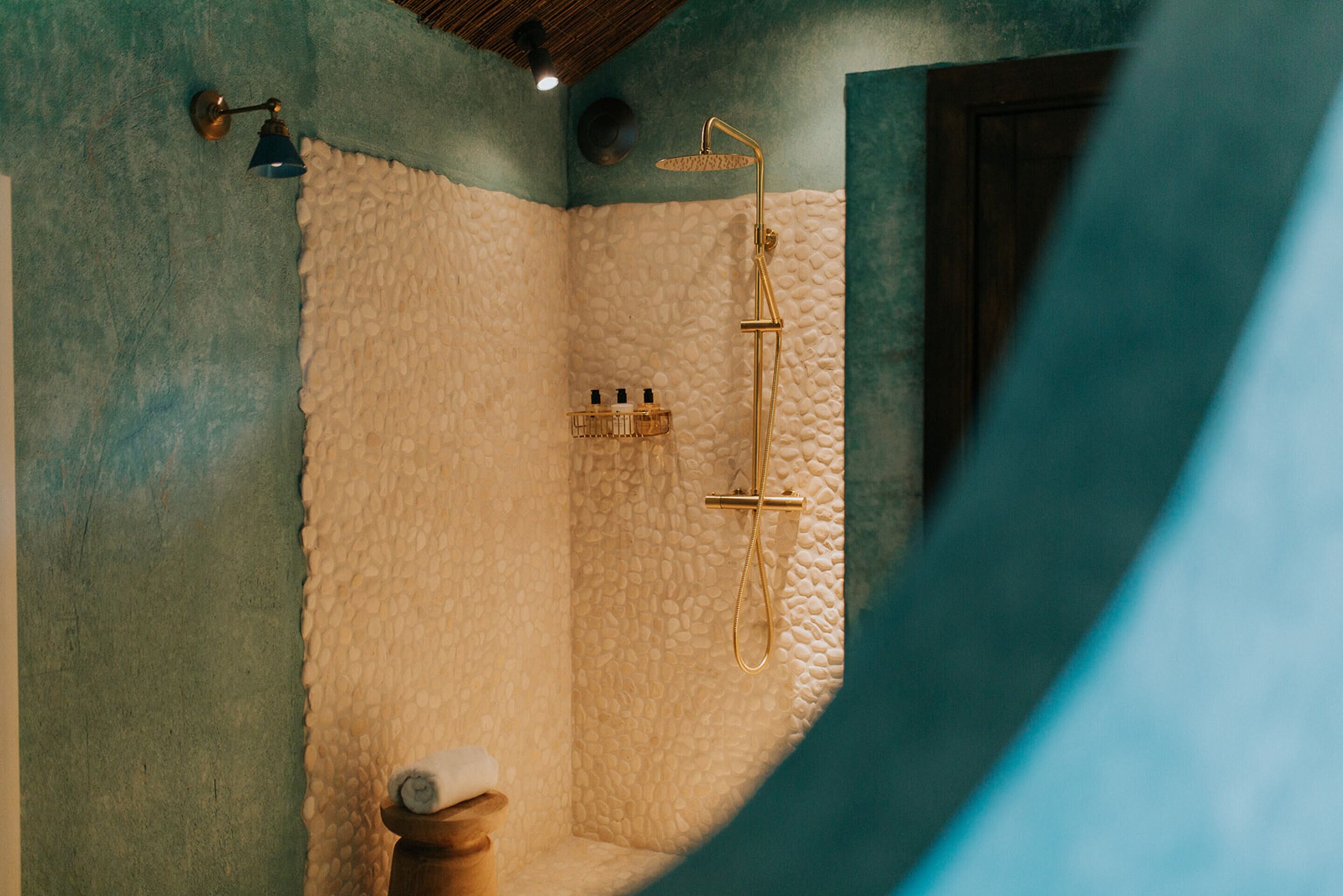 Une salle de bain avec une cabine de douche à l’aspect pierre, une serviette posée sur un tabouret en bois et un luminaire fixé au mur.