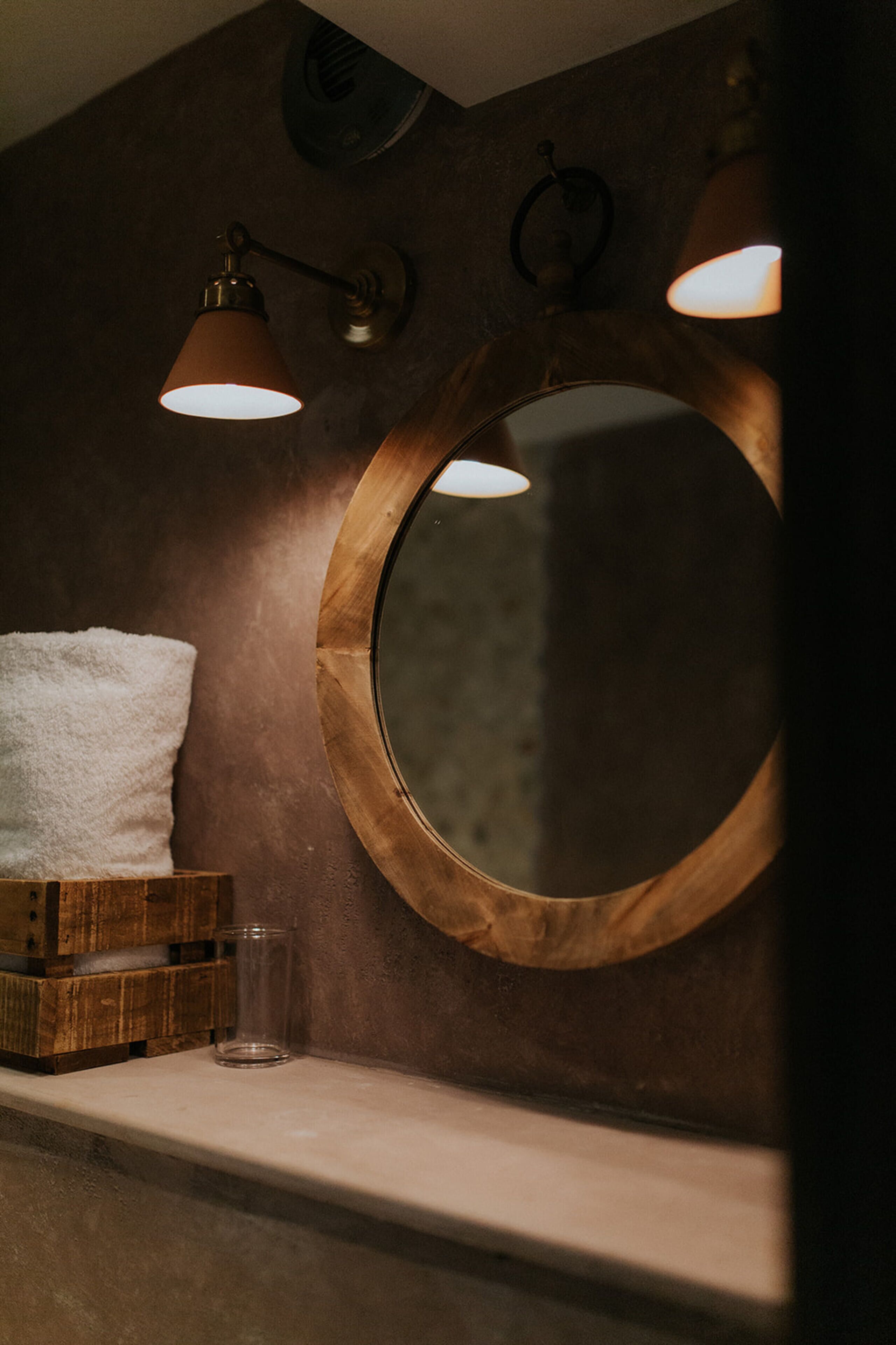 Un baño con un espejo de marco de madera, una toalla blanca y un vaso sobre una estantería de madera.