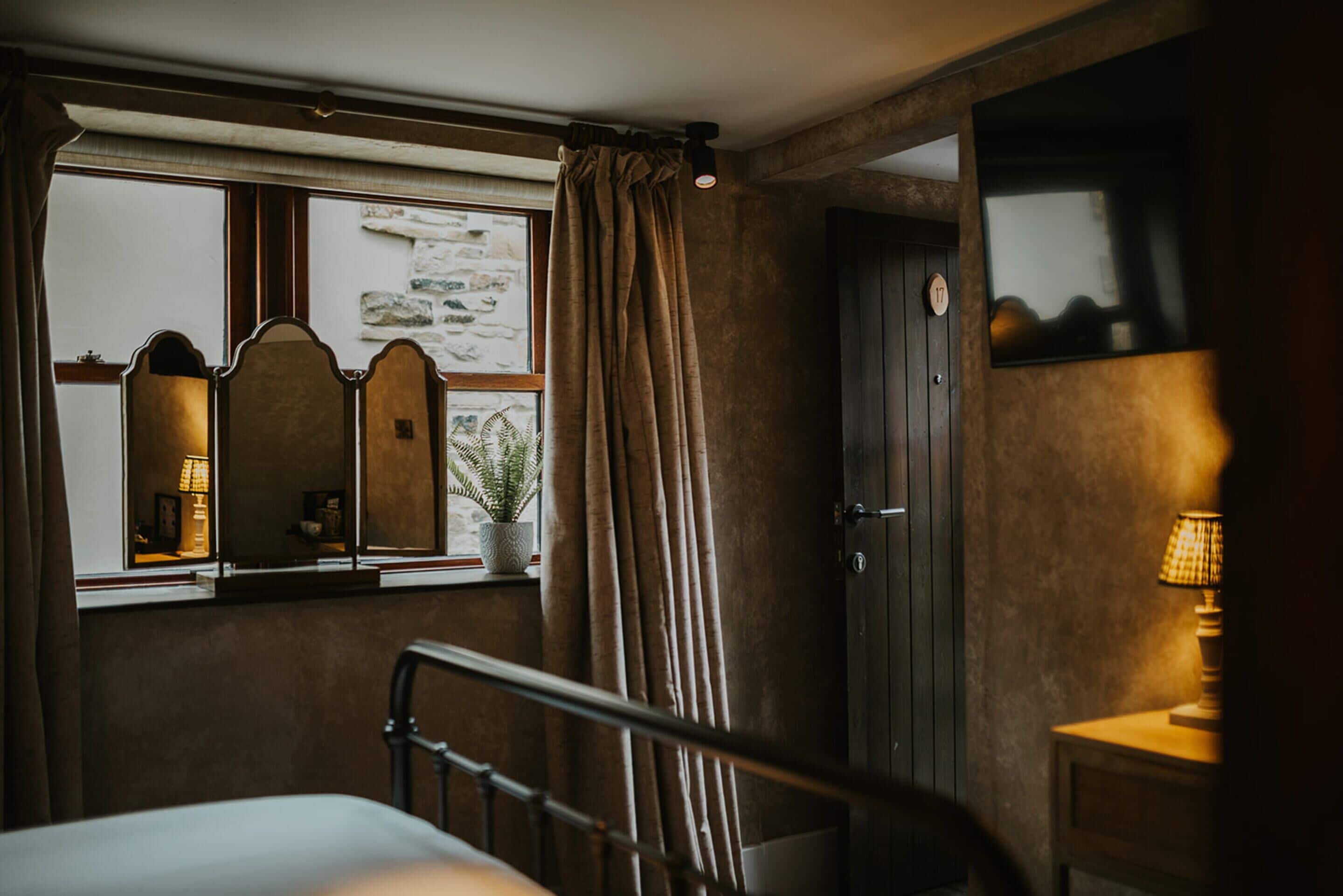 Une chambre à coucher avec un lit, une fenêtre avec des rideaux, une table de chevet avec une lampe et un miroir.