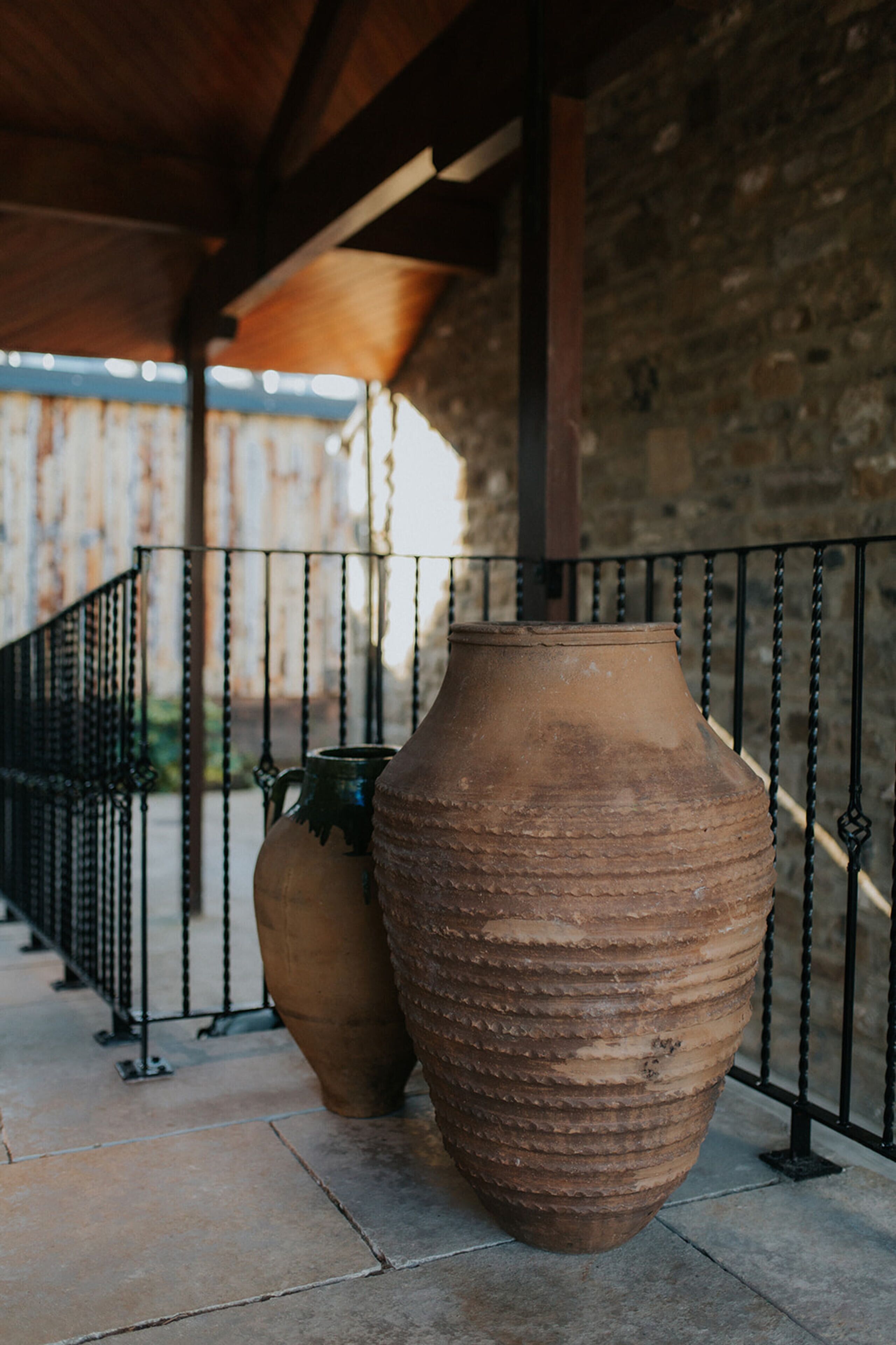Dos grandes macetas de terracota en un patio con baldosas, una barandilla de metal y un muro de piedra al fondo.