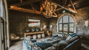 Ein gemütliches Wohnzimmer mit einer großen Blumsofa, einem Holztisch und einem Kronleuchter. Es gibt ein großes Fenster mit Rundbogen, eine Holzbank und einen Korbstuhl.