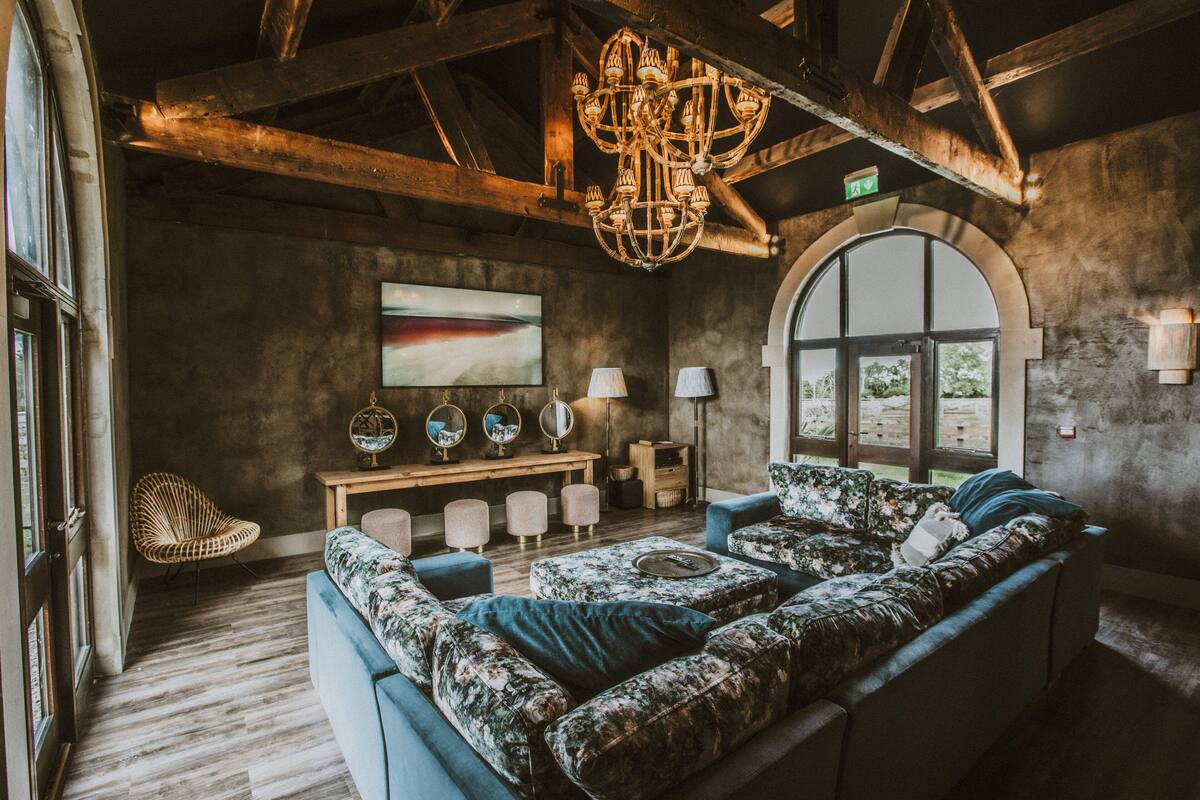 A cozy living room with a large floral sofa, a wooden coffee table, and a chandelier. There is a large window with an arched design, a wooden bench, and a wicker chair.