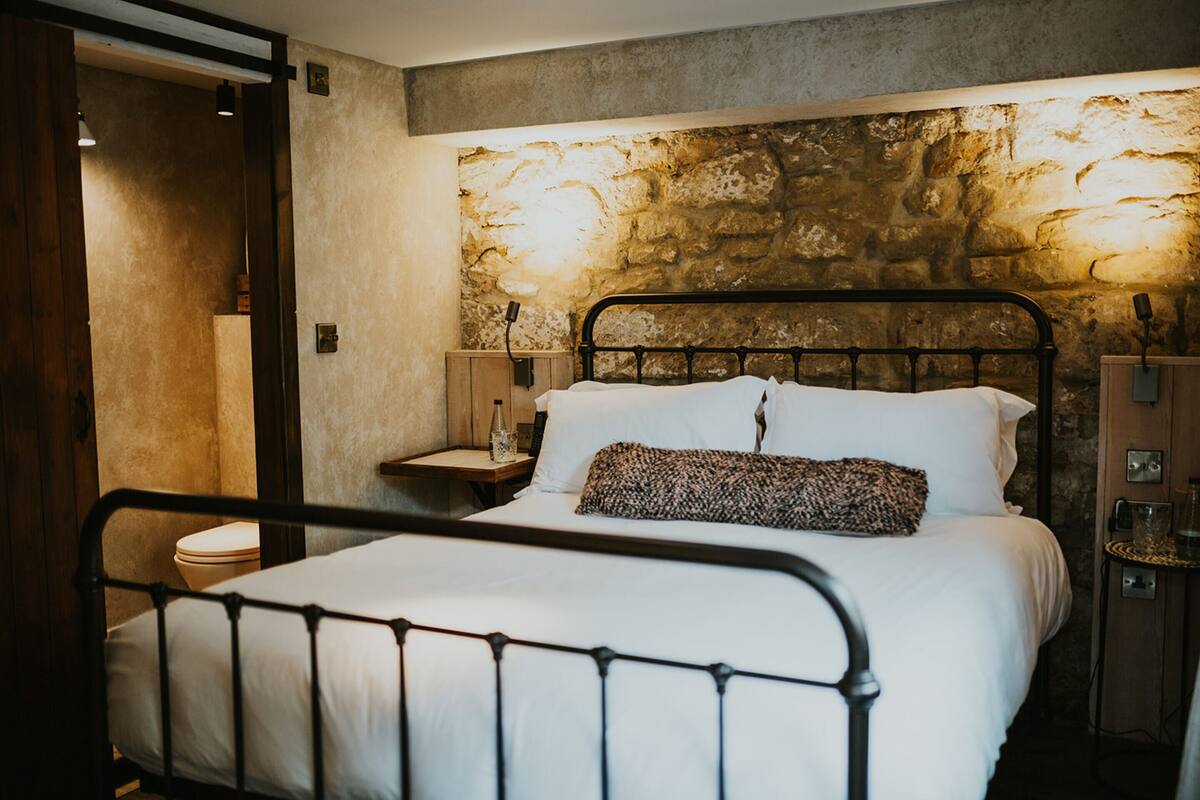 A bed with a metal frame, white bedding, and a patterned pillow. There is a nightstand with a lamp and a wooden cabinet with a mirror. The wall behind the bed is made of stone.