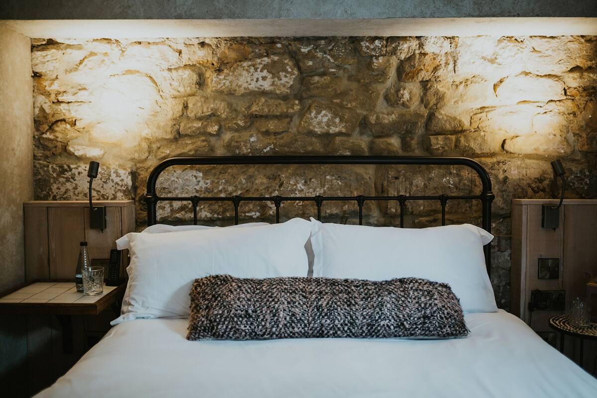 A bed with white linens and a patterned pillow, set against a stone wall backdrop.