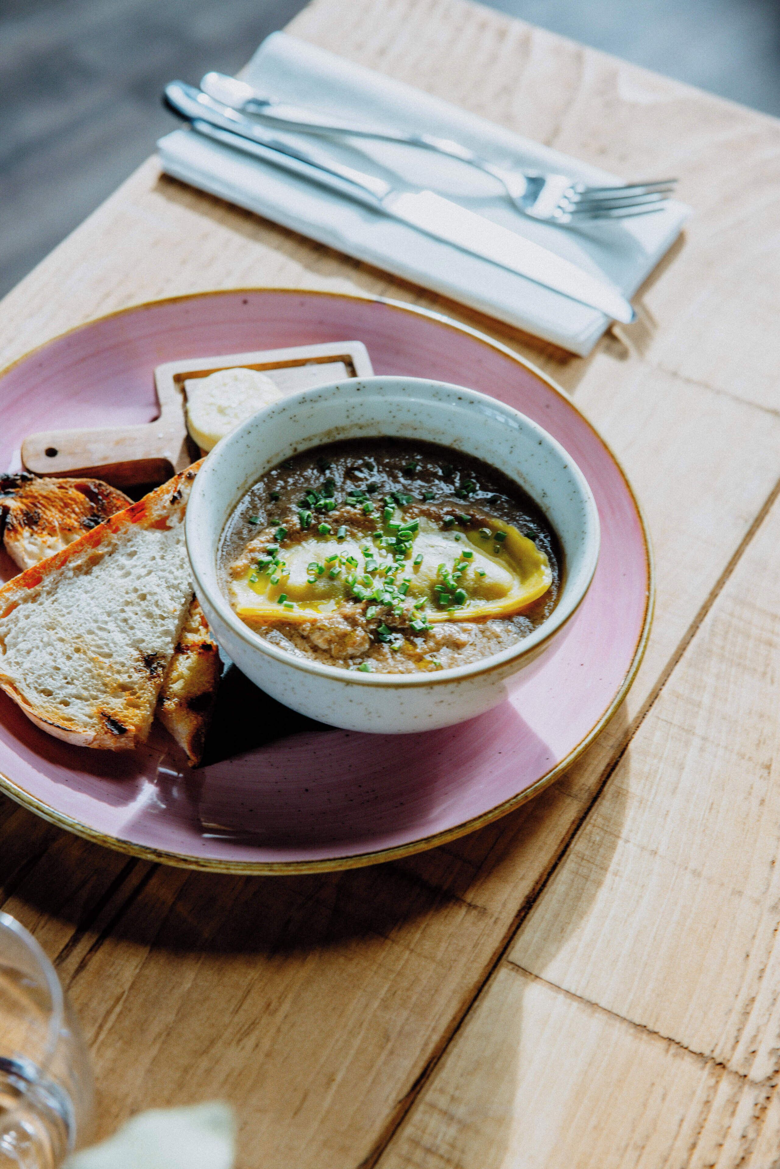 A bowl of soup on a pink plate with toasted bread beside it, placed on a wooden table.