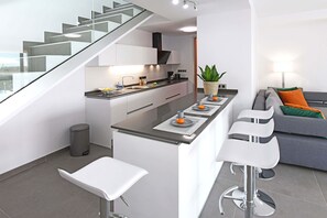 A modern kitchen with a glass staircase, a dining area with bar stools, and a grey sofa with orange cushions. - Salobre La Isla 8 (Maspalomas)