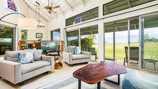 A spacious living room with a grey sofa, a coffee table, and a TV. There is a large window with a view of the outdoors.