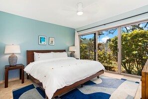 A bedroom with a large bed, two bedside tables, a ceiling fan, and a view through a sliding glass door to an outdoor area with plants.