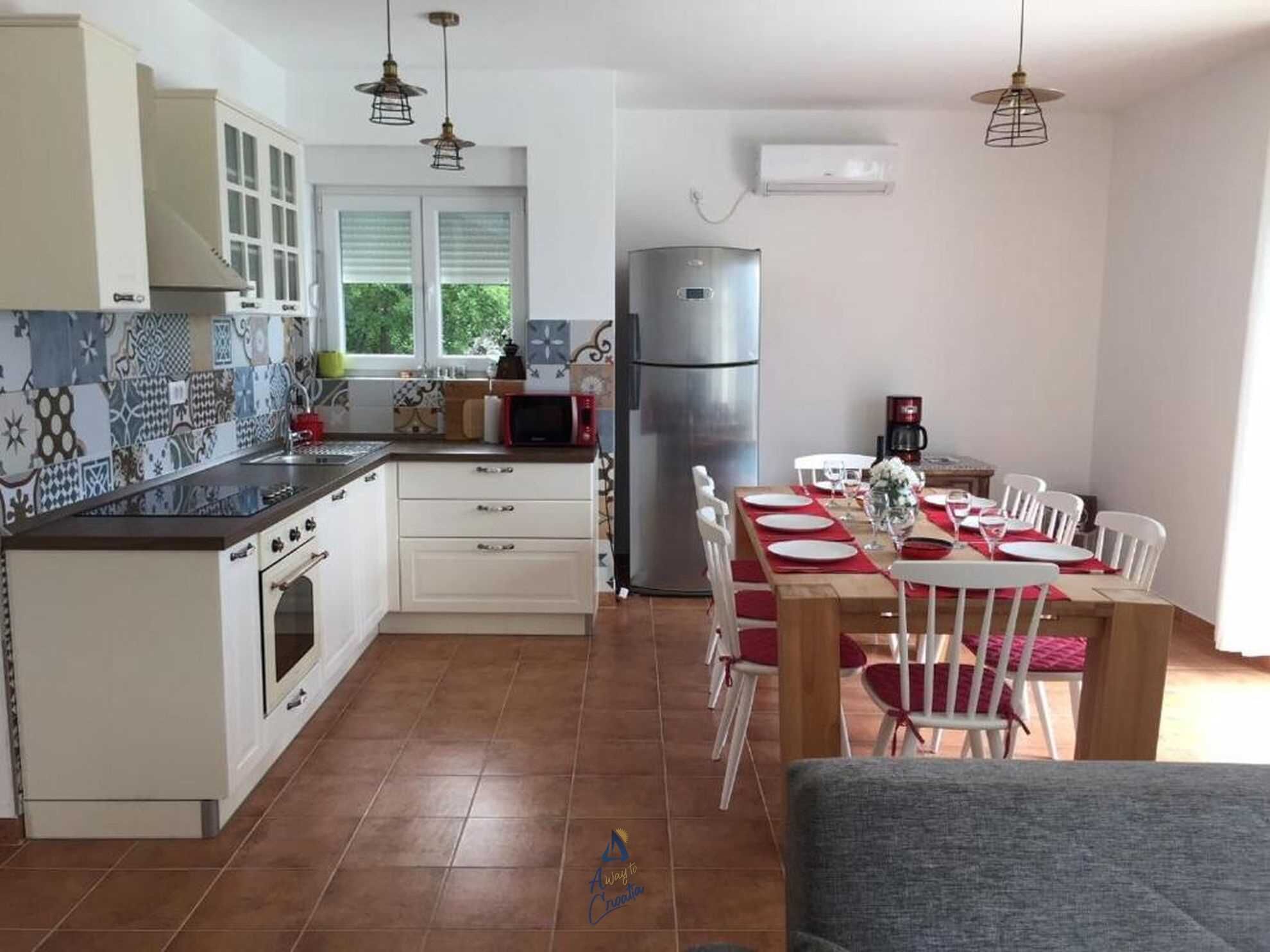 A kitchen with white cabinets, a stainless steel refrigerator, a dining table with white chairs, and hanging pendant lights.