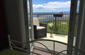 A balcony with a wicker chair, a small table with flowers, and a view of a body of water and greenery.
