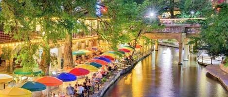 A riverside walkway with outdoor seating under umbrellas, a bridge, and buildings in the background.
