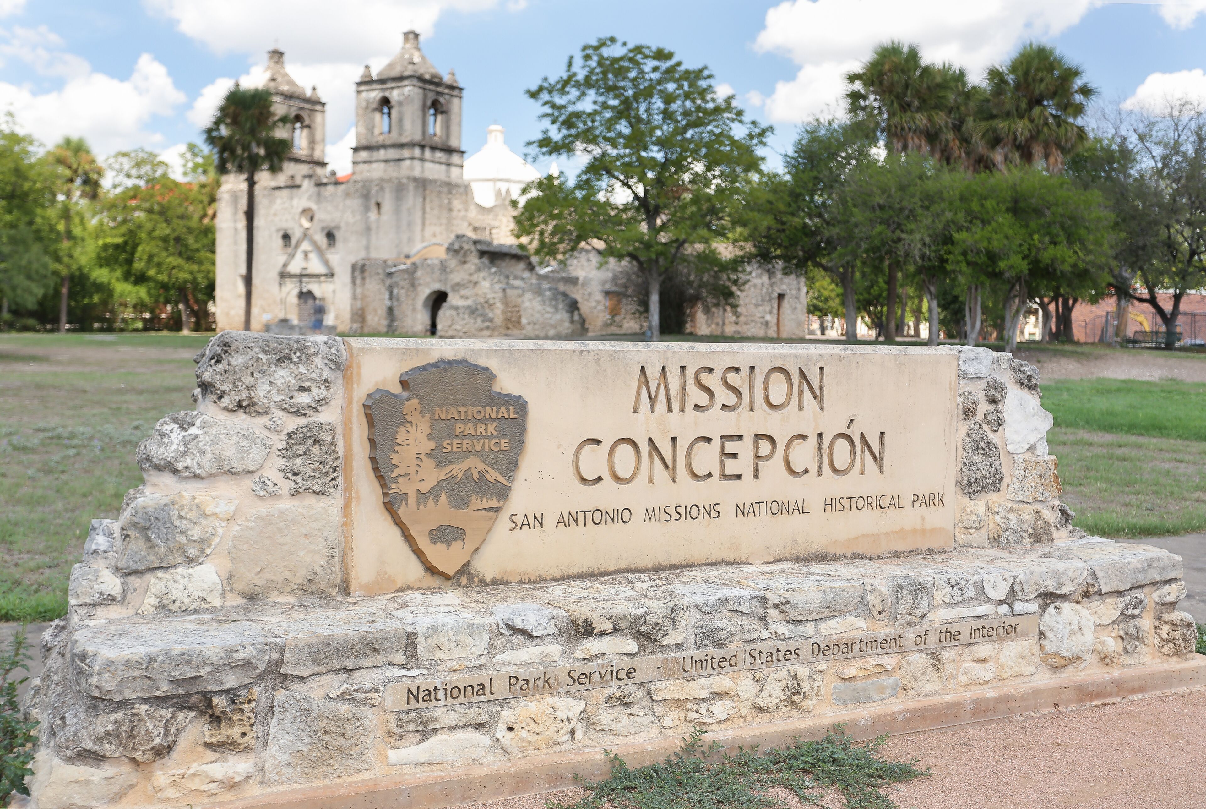 A historical park sign with 'MISSION CONCEPCION' and 'SAN ANTONIO MISSIONS NATIONAL HISTORICAL PARK' inscribed.