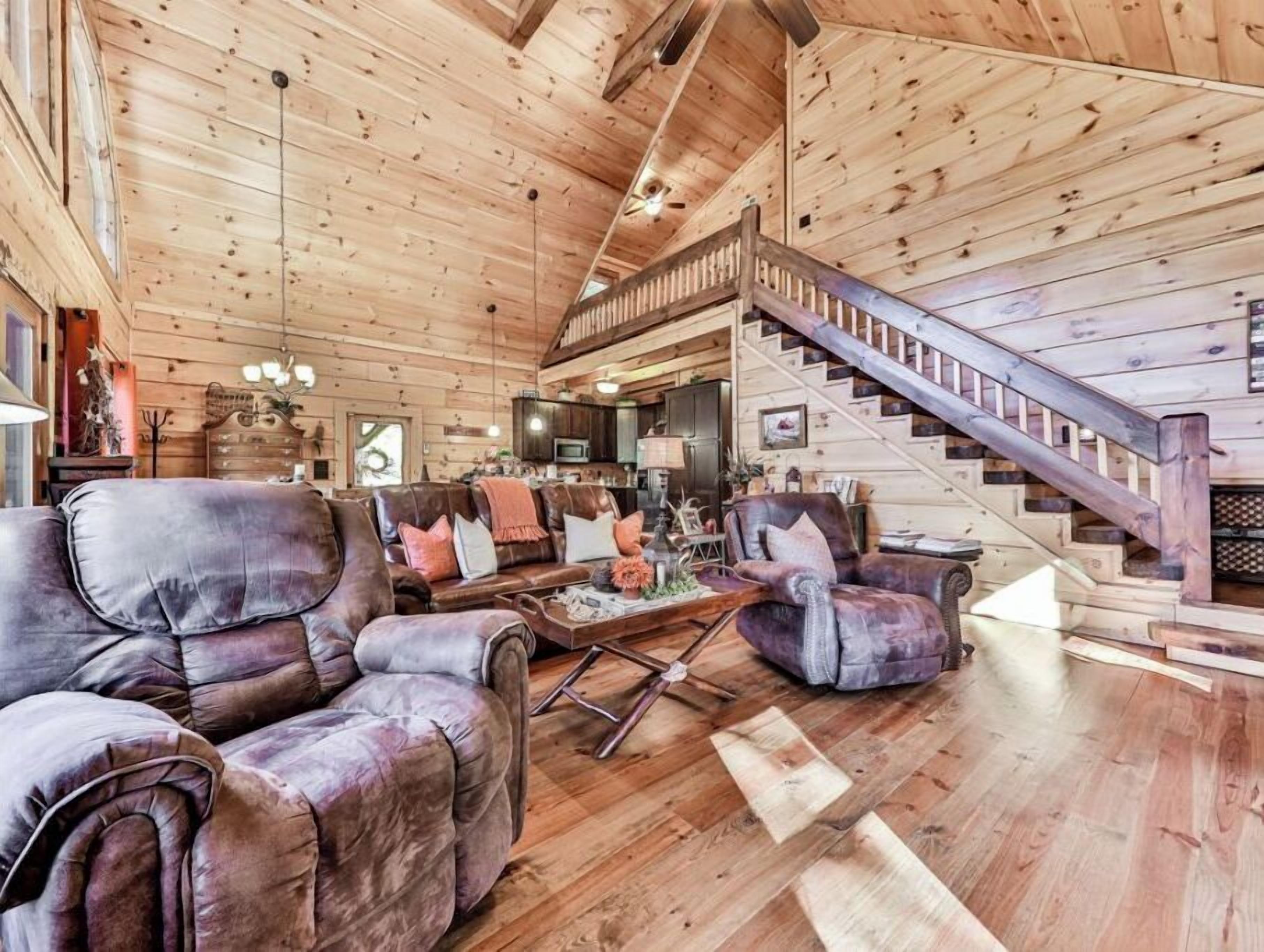 A spacious living room with leather sofas, a wooden staircase, and a kitchen in the background.