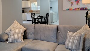 Un salón con un sofá gris, cojines a rayas, un ventilador de techo y una cocina al fondo.