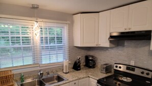 Una cocina con armarios blancos, un lavamanos de acero inoxidable, una estufa y una ventana con persianas.