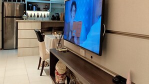 A modern living room with a flat-screen TV mounted on the wall, a kitchen in the background, and a dining area with a white table and chairs. - Nautilus Concept - Penha-SC (Penha)