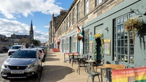 A street view with parked cars, outdoor seating at a cafe, and a building with a sign for 'Passey's'. - Immaculate Family sized holiday home located 10mins from beachfront! (Edinburgh)