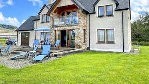 Une maison à deux étages avec une façade en pierre, un balcon et un patio aménagé avec du mobilier d’extérieur.