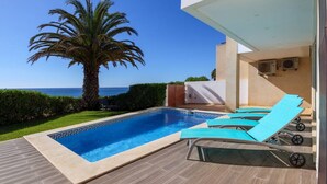 Un espace aménagé au bord de la piscine, offrant une vue dégagée sur l’océan, avec un palmier et un bâtiment moderne doté d’un patio couvert.