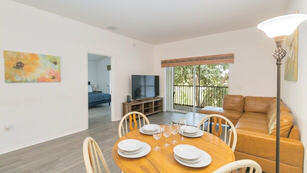 A dining area with a wooden table set for four, a television in the background, and a sliding glass door leading to a balcony. - Huge screened patio that overlooks Lake Buck, near famous attractions!! (Kissimmee)
