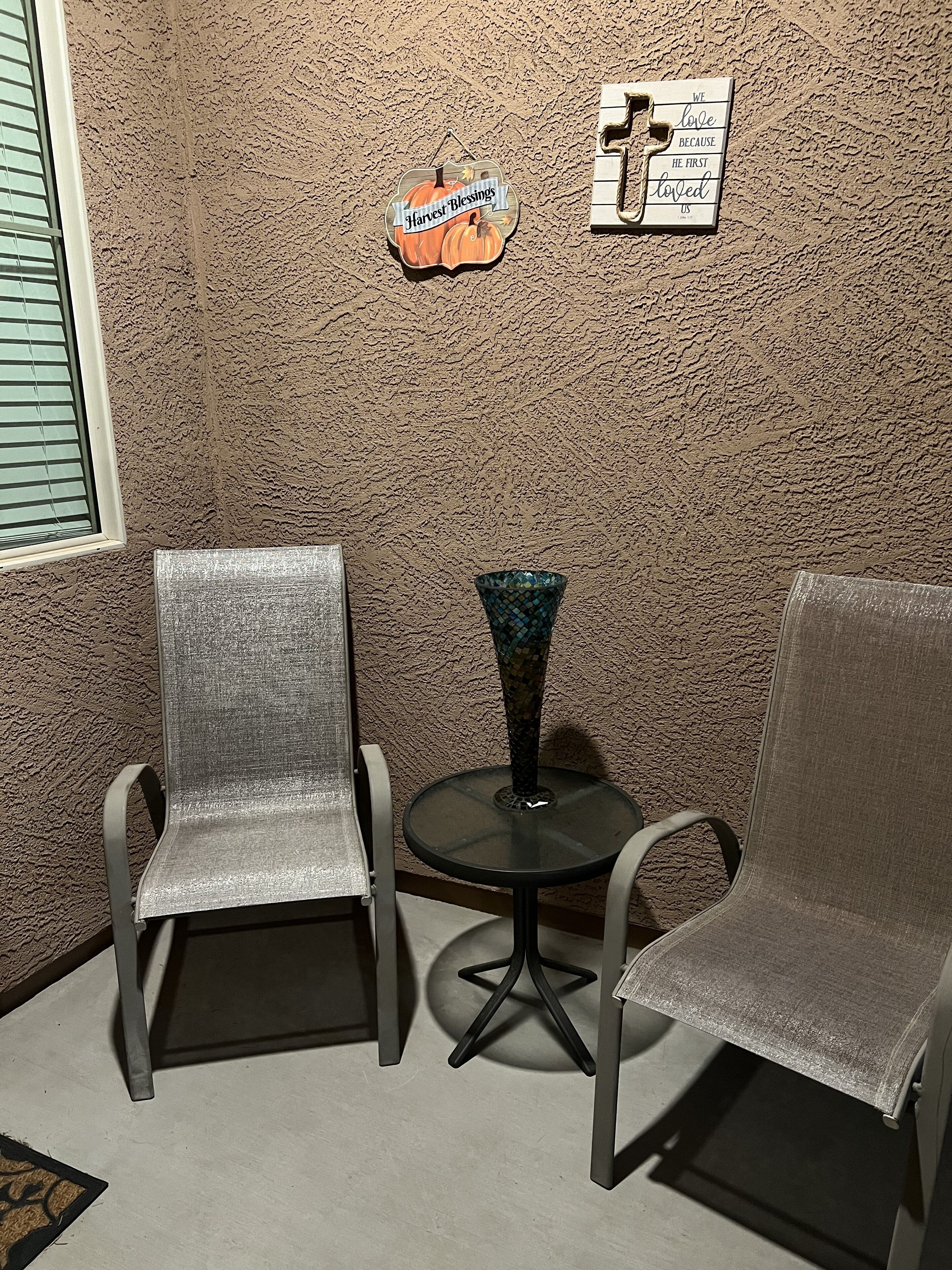 Deux chaises en métal, une petite table et un vase décoratif placés entre elles, le tout adossé à un mur texturé orné d’une pancarte indiquant « Bénédictions de la récolte » et d’une autre pancarte indiquant « Nous aimons parce qu’il nous a aimés en premier ».