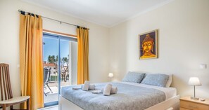 Quarto com cama, mesa de cabeceira, cadeira, janela com cortinas e um quadro na parede.