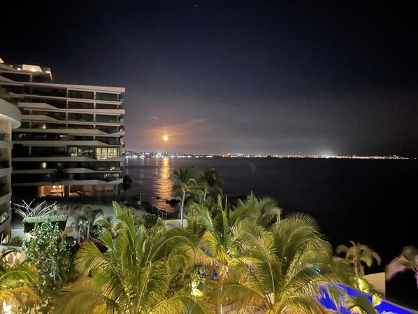 Vue nocturne d’un hôtel situé sur la côte, avec un balcon donnant sur la mer, des palmiers et un paysage urbain lointain.